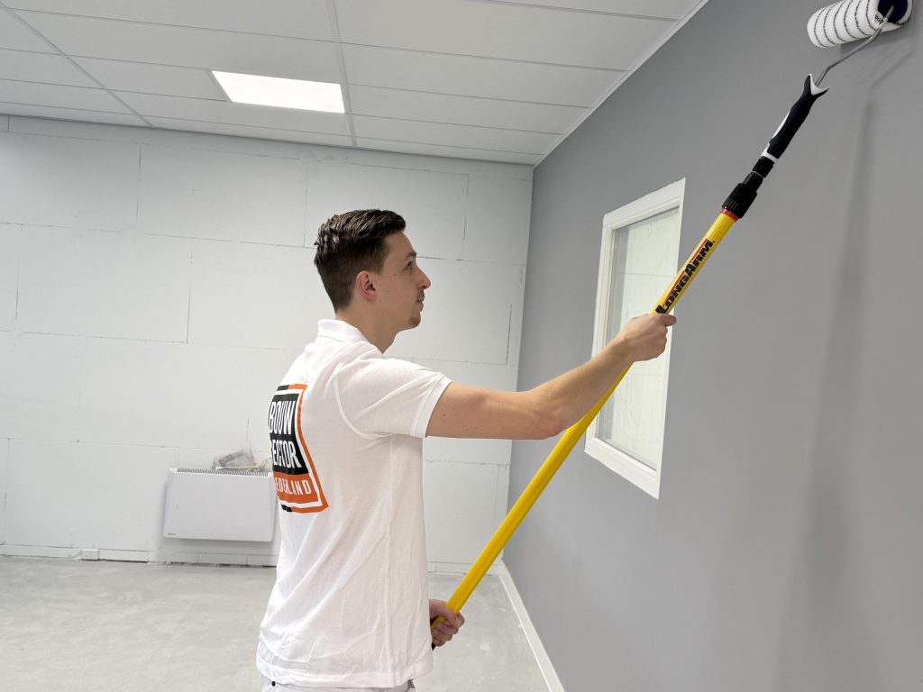 Schilder werkt met een lange roller langs een wit raamkozijn in een grijze wand