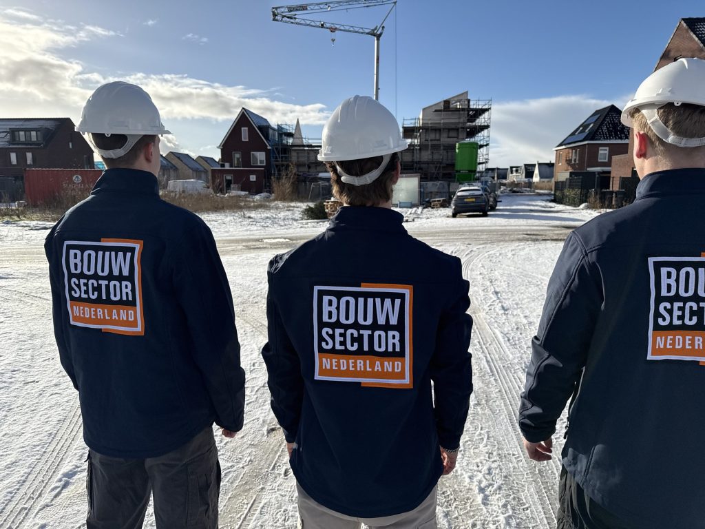 Drie vakmensen van Bouwsector Nederland met witte helm kijken uit over een woonwijk in aanbouw
