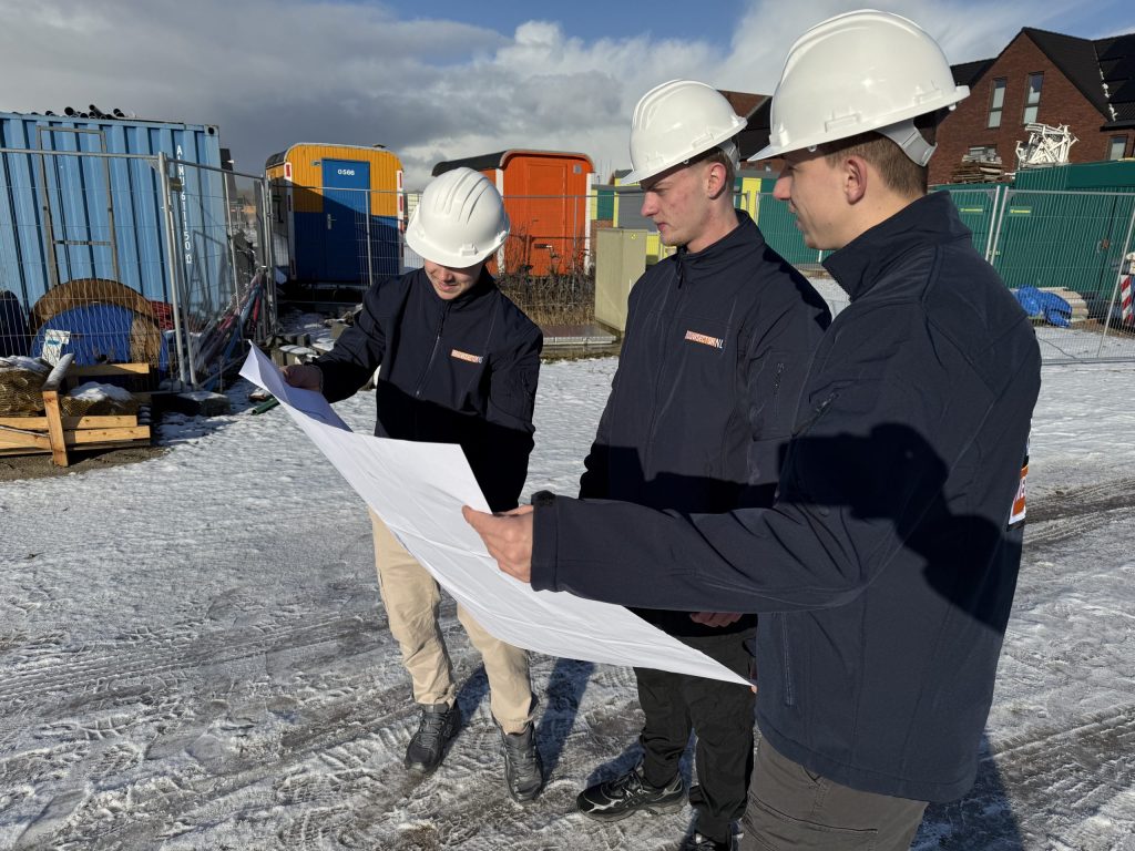 Drie vakmensen van Bouwsector Nederland met witte helm bekijken samen een bouwtekening op een besneeuwd terrein