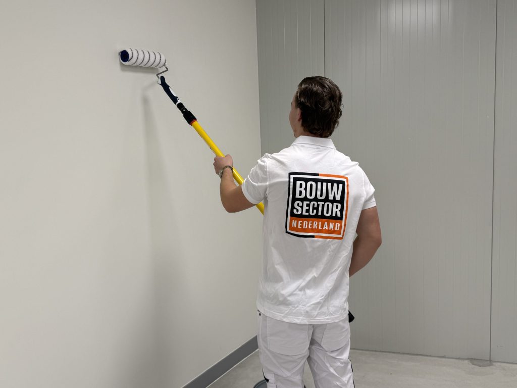 Schilder van Bouwsector Nederland rolt een witte wand in een lege ruimte met een lange roller