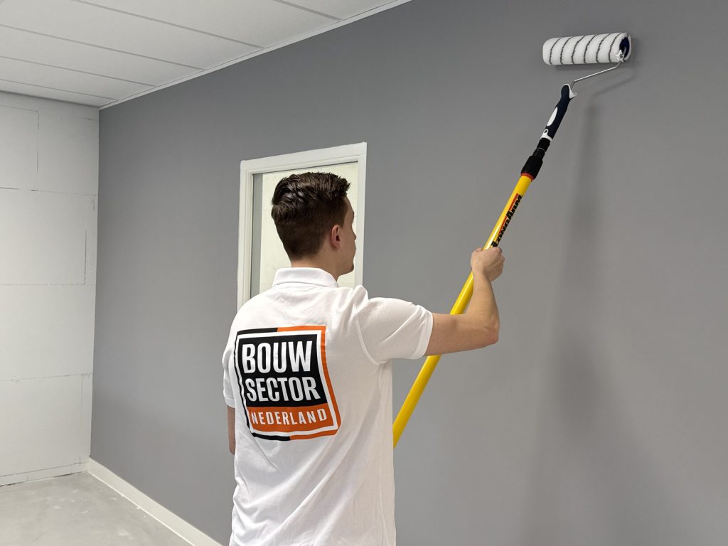 Schilder van Bouwsector Nederland rolt een grijze wand met een roller aan een gele verlengsteel, onder een plafond van platen